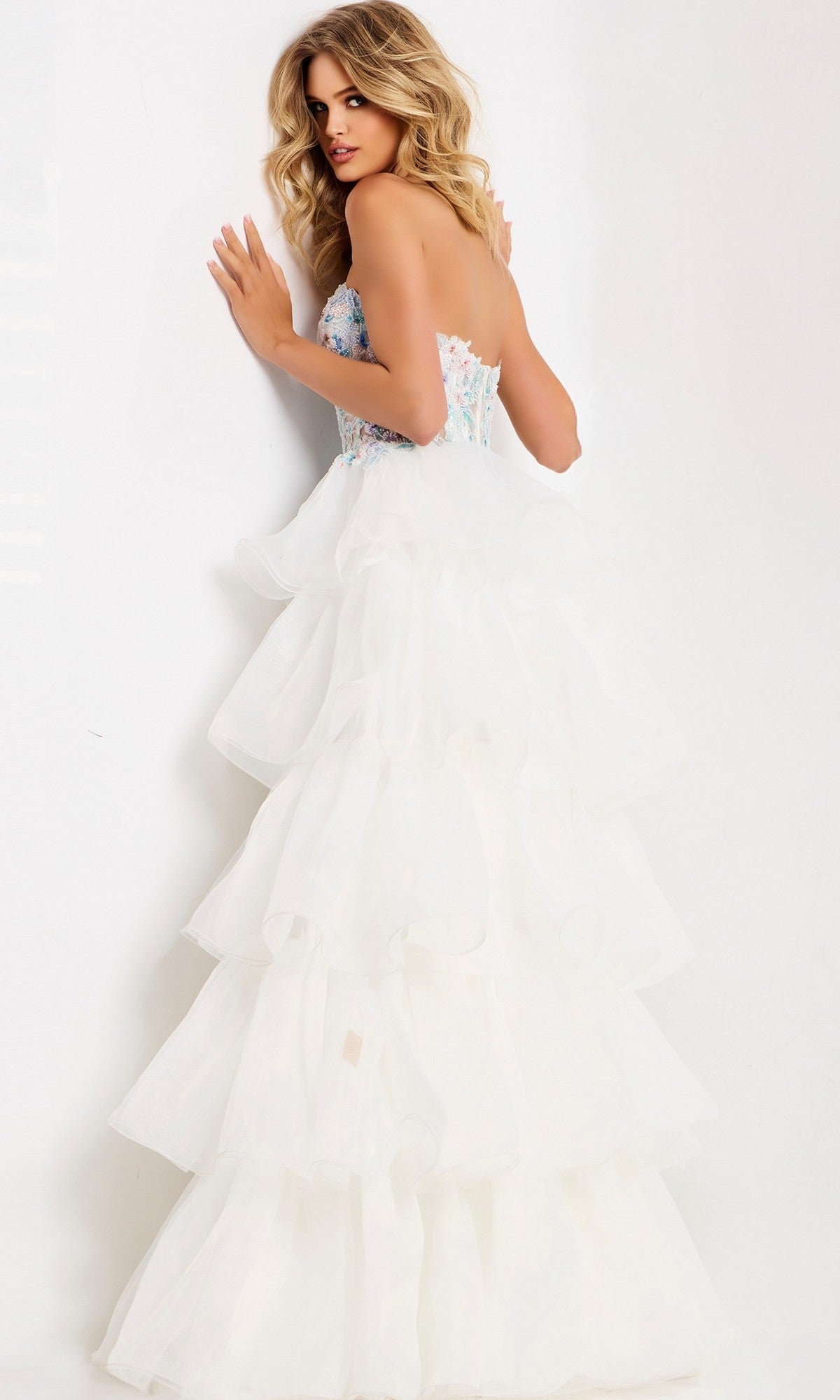 A woman with wavy blonde hair wears the Jovani 48177 long prom dress, a strapless white gown with a fitted floral and beaded corset bodice, standing by a white wall and looking over her shoulder.